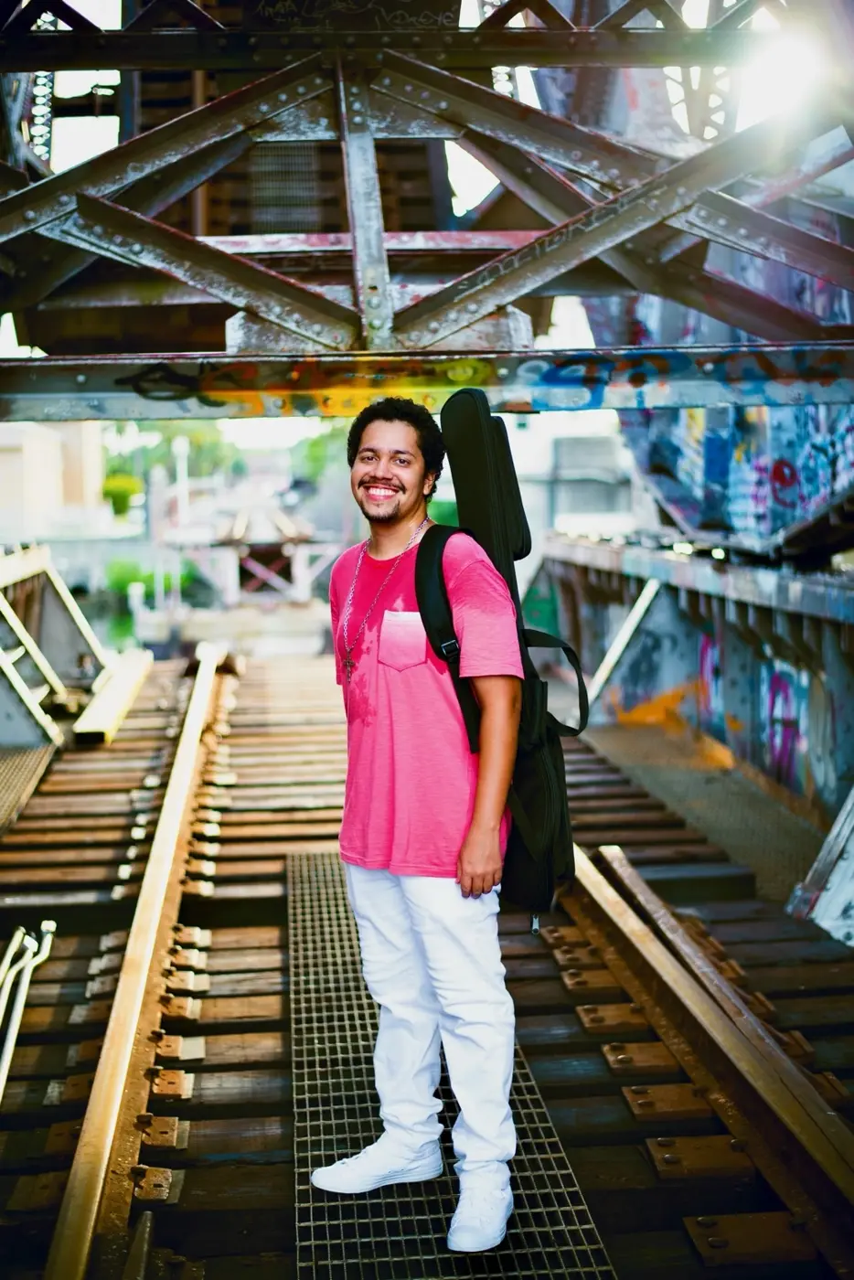 Smiling musician in a pink shirt carrying a guitar case beneath a steel bridge