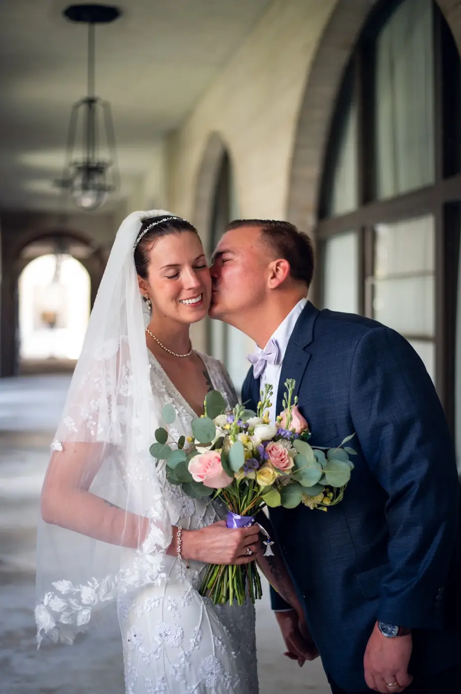 Groom kissing the bride on the cheek beneath a stone arcade