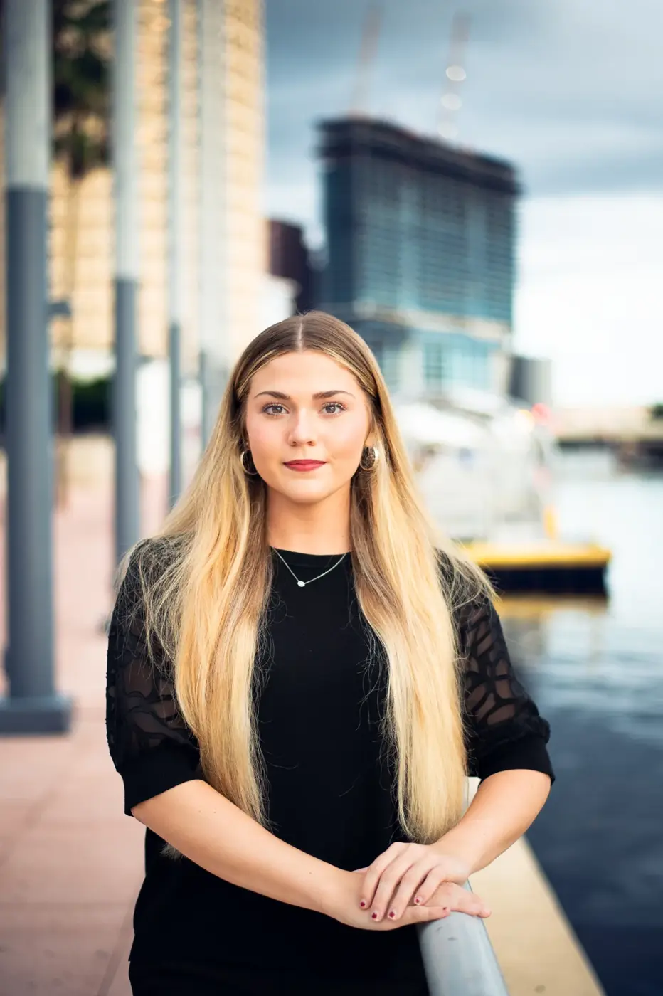 Portrait of a woman in black beside the waterfront, with city buildings softly blurred behind her